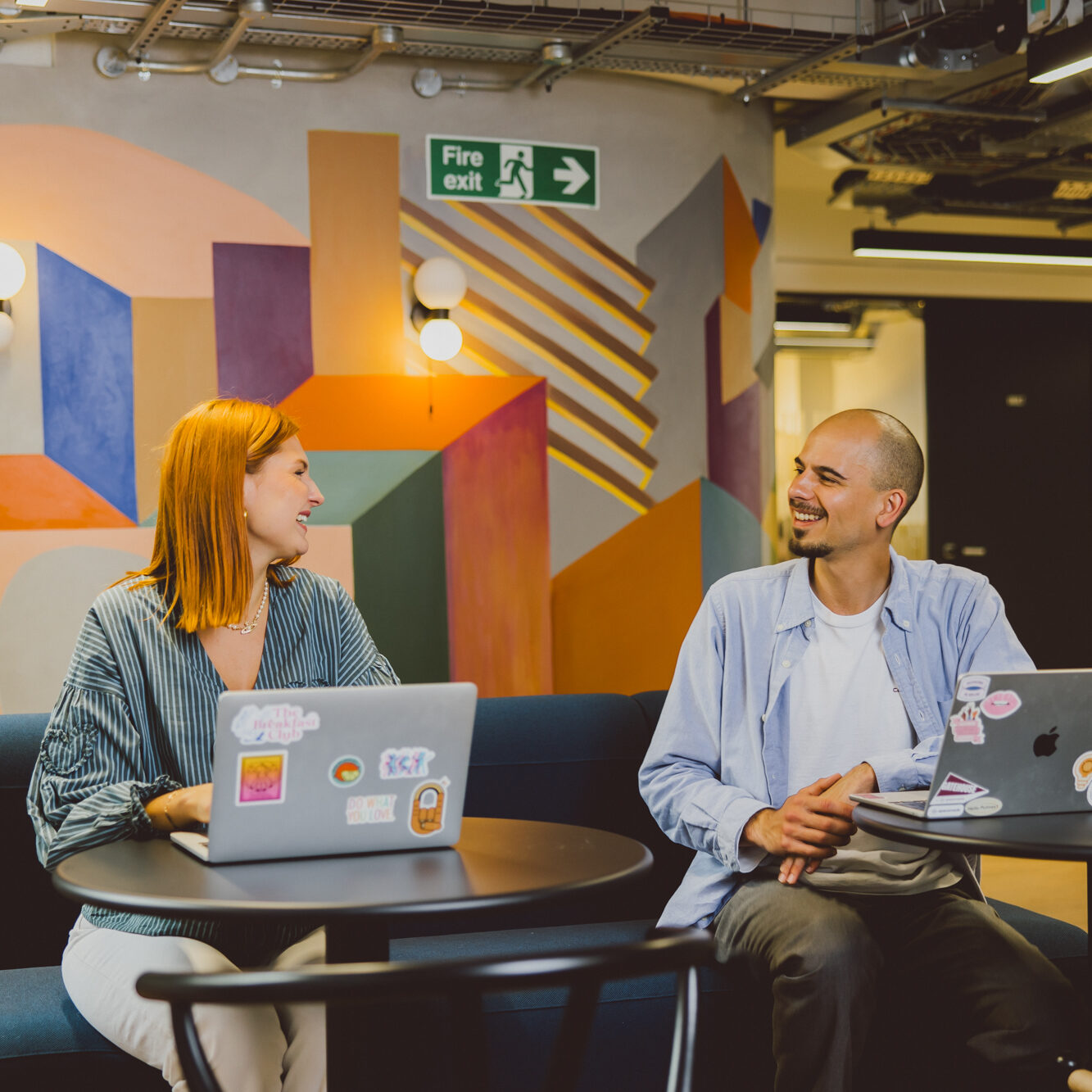 Two people smiling and chatting while working on laptops in a vibrant coworking space with colourful mural walls and a relaxed, creative atmosphere. Two people smiling and chatting while working on laptops in a vibrant coworking space with colourful mural walls and a relaxed, creative atmosphere.