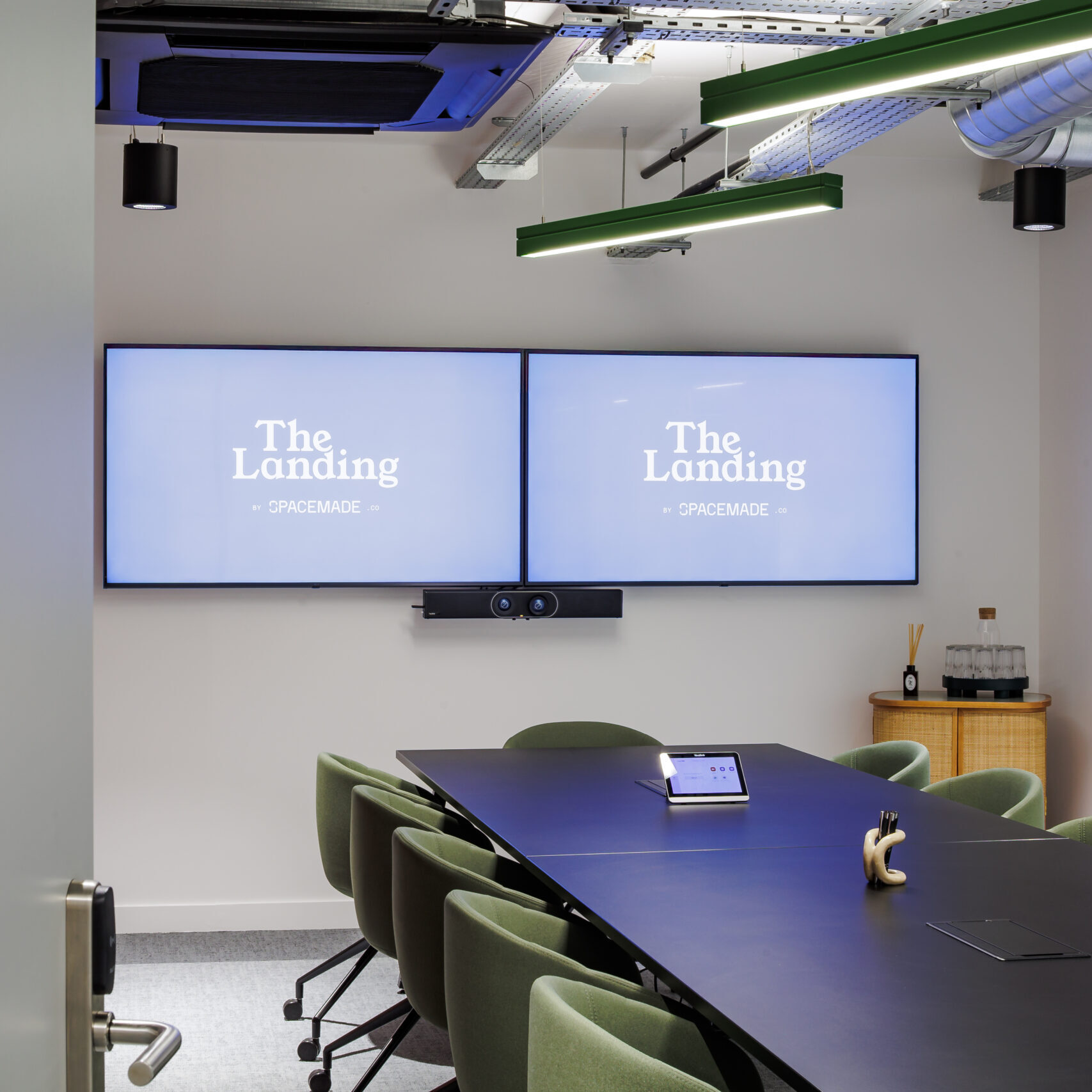Spacious 12-person meeting room in a coworking space in Putney, featuring dual screens, a sleek black table, green upholstered chairs, and a fully equipped video conferencing setup.