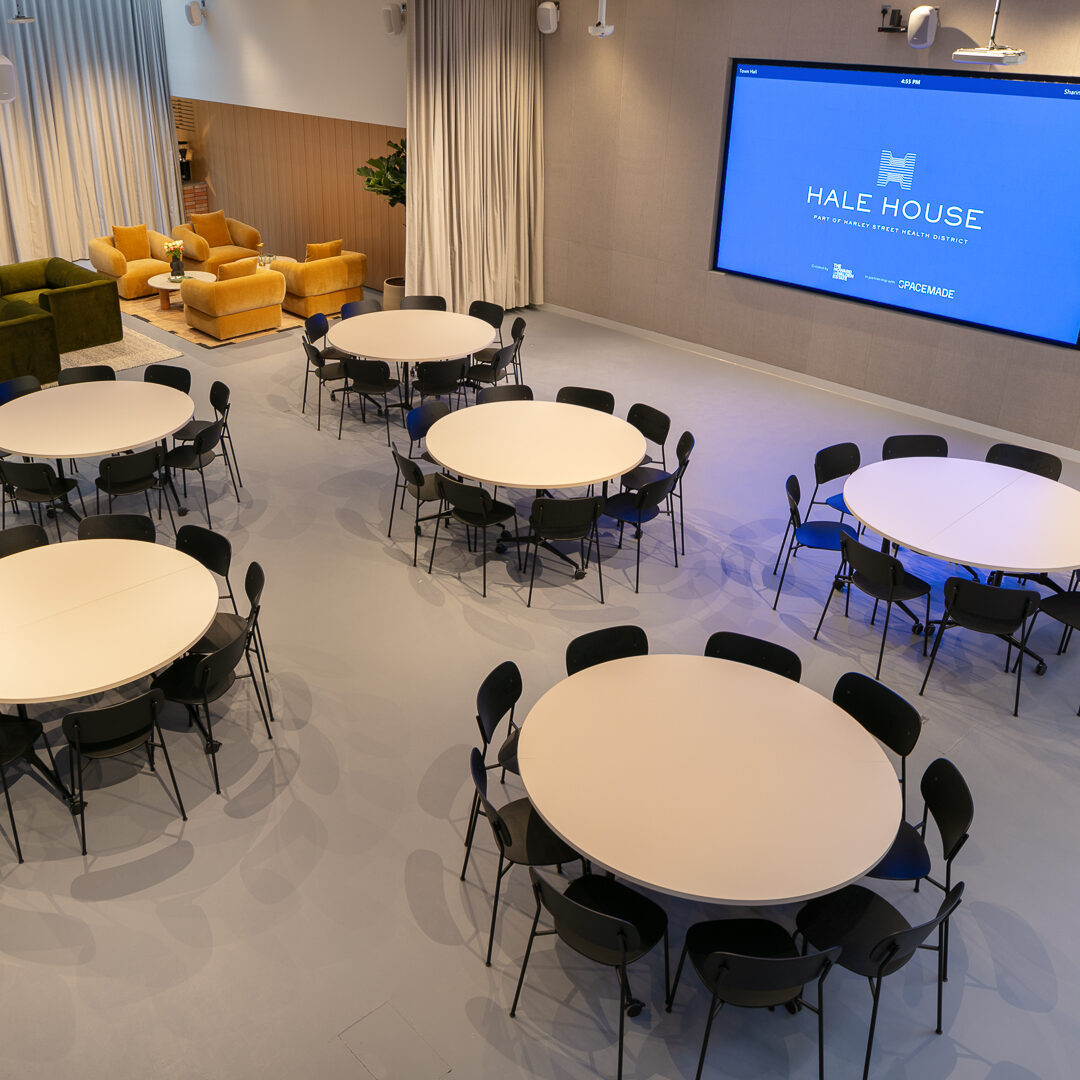 Versatile event space at Hale House set up with round tables and chairs for a conference or workshop, featuring a large presentation screen and stylish lounge area in the background.