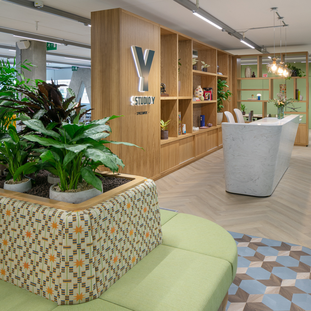 Modern and vibrant reception area at Studio Y coworking space in Notting Hill, featuring lush indoor plants, a patterned bench, geometric flooring, and a sleek white marble desk set against warm wooden shelving.