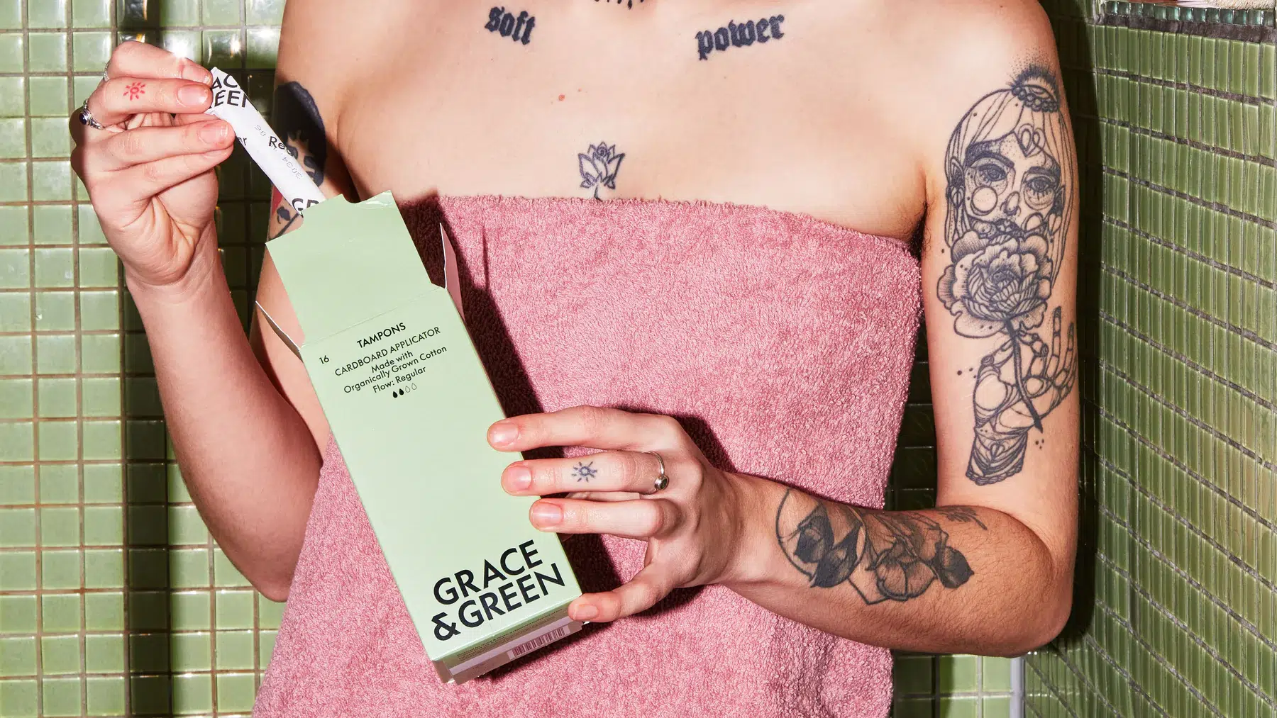 Person with visible tattoos and a towel wrapped around their body holding a Grace & Green box of organic period products in a tiled bathroom setting. Person with visible tattoos and a towel wrapped around their body holding a Grace & Green box of organic period products in a tiled bathroom setting.