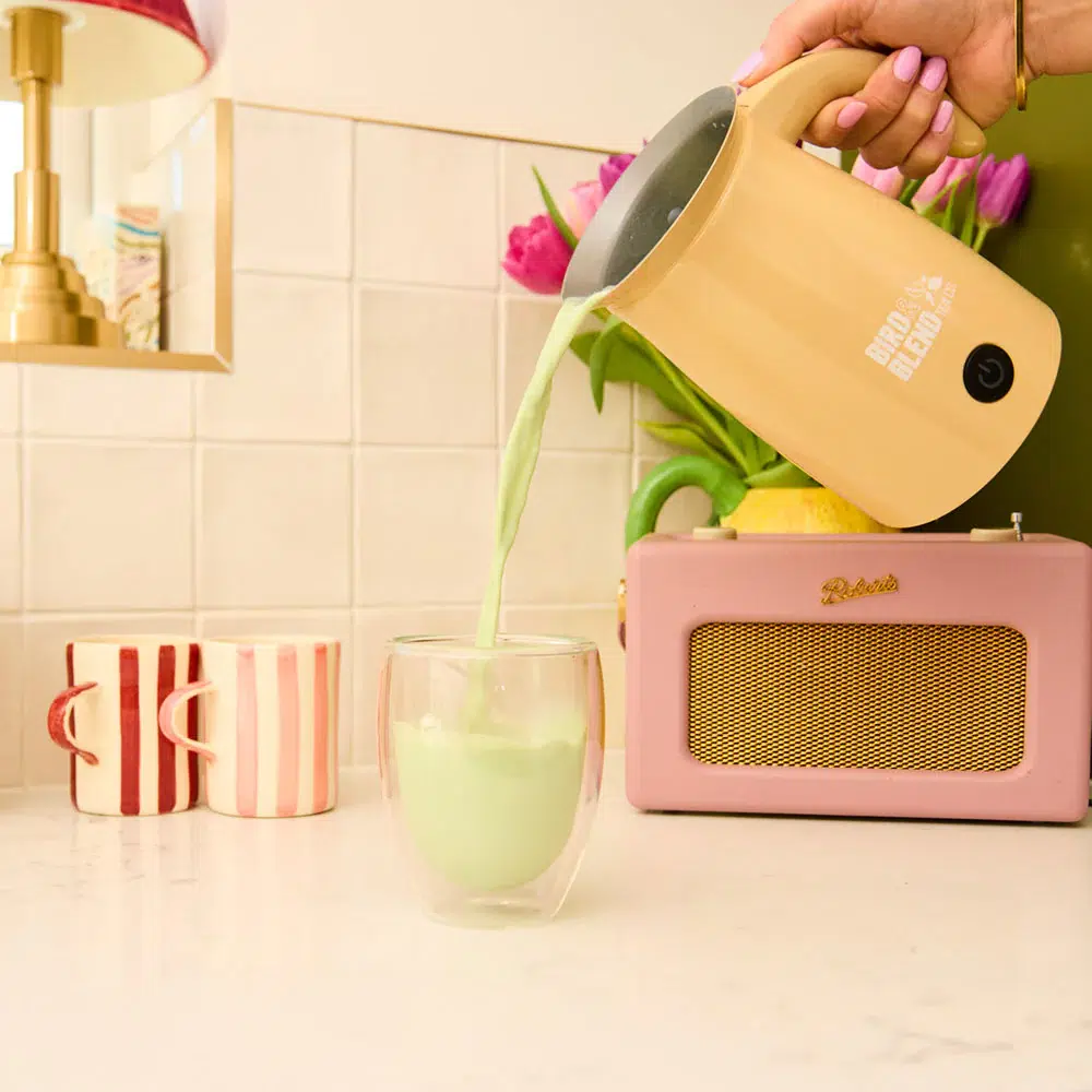 A hand pours a vibrant green matcha latte from a yellow Bad Blend frother into a glass mug on a pastel-themed kitchen counter, with striped mugs, a pink Roberts radio, and tulips in the background. A hand pours a vibrant green matcha latte from a yellow Bad Blend frother into a glass mug on a pastel-themed kitchen counter, with striped mugs, a pink Roberts radio, and tulips in the background.