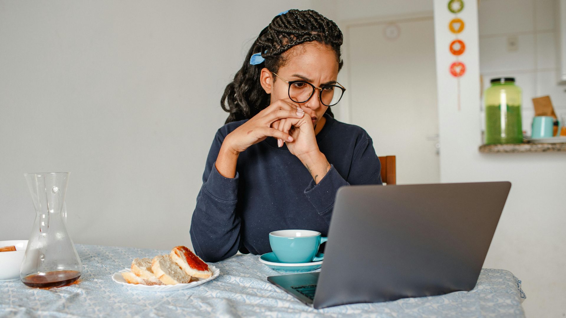 A woman sitting at her kitchen table, frowning whilst looking at her laptop with headphones on. There is a blue tea cup and a plate of bread on the table.